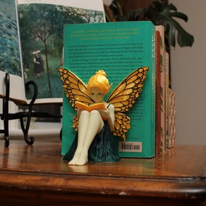 May include: A fairy ornament with golden wings and a yellow head is reading a book. The fairy is positioned in front of a teal book. Other books are visible in the background, and the ornament is on a wooden surface.