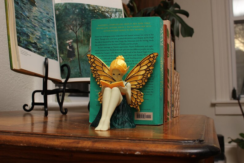 May include: A fairy ornament with golden wings and a yellow head is reading a book. The fairy is positioned in front of a teal book. Other books are visible in the background, and the ornament is on a wooden surface.