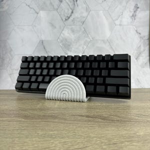 May include: A white, curved keyboard stand with a black keyboard resting on top. The stand is made of a textured material and has a rounded shape.