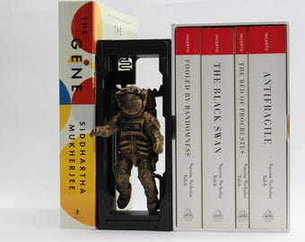 May include: A bronze astronaut figurine stands in a black frame between several books. The books have white covers with red accents and titles like "ANTIFRAGILE" and "THE BLACK SWAN." One book has a colorful cover with the title "THE GENE."