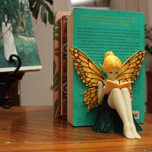 May include: A decorative fairy figurine with yellow hair and butterfly wings, reading a book, sits in front of a stack of books. The fairy is wearing a green dress. The books are on a wooden table.