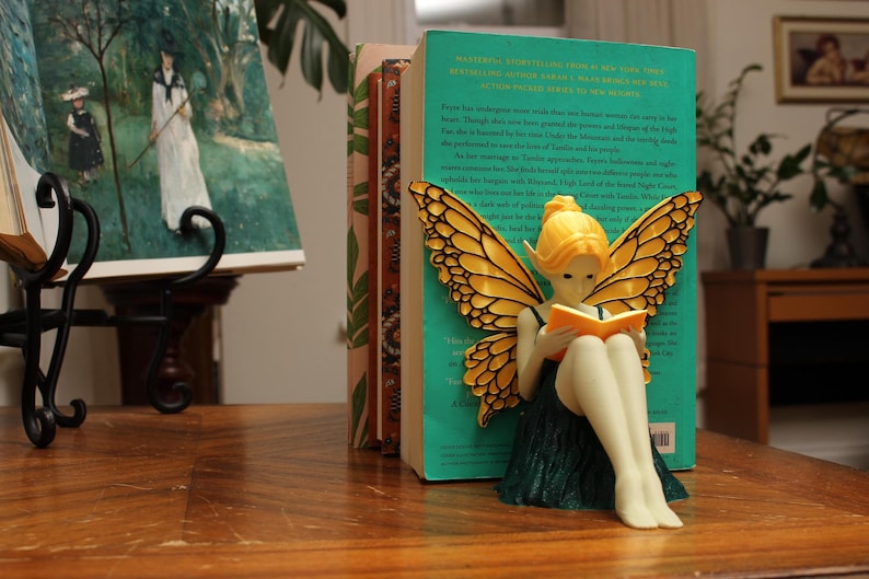 May include: A decorative fairy figurine with yellow hair and butterfly wings, reading a book, sits in front of a stack of books. The fairy is wearing a green dress. The books are on a wooden table.