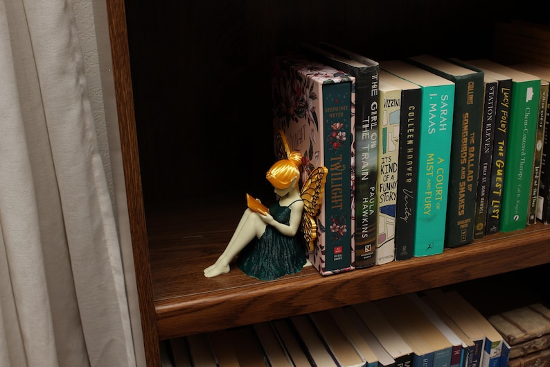 May include: A fairy figurine with golden wings and hair, wearing a dark green dress, reading a book. The figurine sits on a wooden shelf beside a row of books with colourful spines, including "Twilight" and "The Guest List".