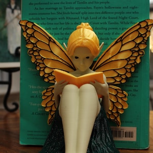 May include: A fairy figurine with orange wings and hair, reading a book. The fairy is seated with its legs crossed, wearing a dark green dress. The wings have a detailed pattern. A book is propped up behind the fairy.