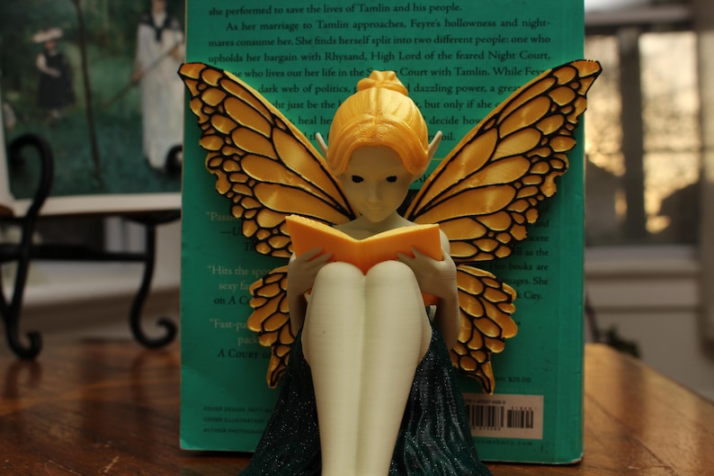 May include: A fairy figurine with orange wings and hair, reading a book. The fairy is seated with its legs crossed, wearing a dark green dress. The wings have a detailed pattern. A book is propped up behind the fairy.