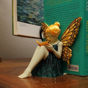 May include: A fairy figurine with golden wings and hair, dressed in a dark green gown, reading an orange book. The fairy is placed beside a stack of books, including "A Court of Mist and Fury".