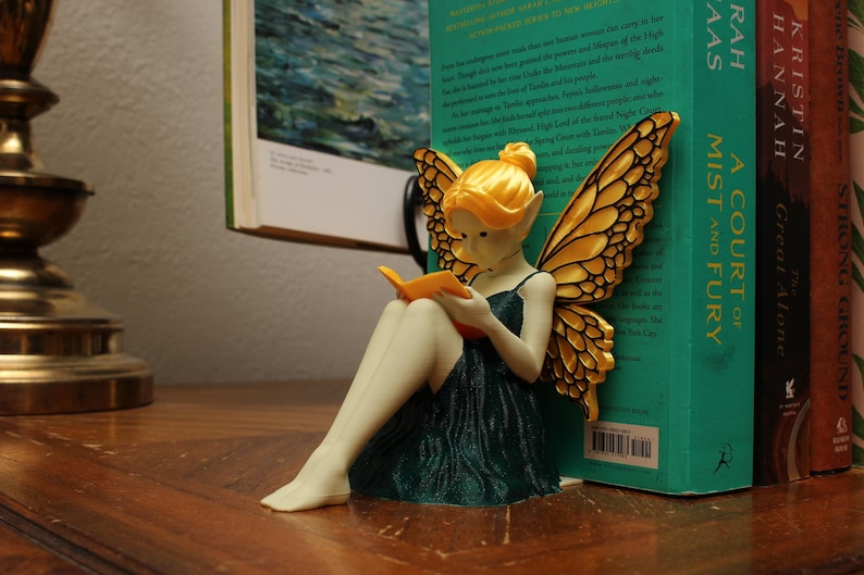 May include: A fairy figurine with golden wings and hair, dressed in a dark green gown, reading an orange book. The fairy is placed beside a stack of books, including "A Court of Mist and Fury".