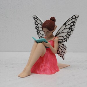 May include: A fairy figurine with brown hair and intricate wings. The fairy is dressed in a pink dress and reading a small, blue book. The fairy is seated with her legs crossed.