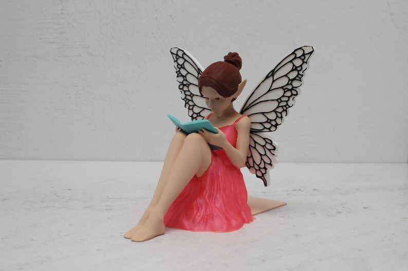 May include: A fairy figurine with brown hair and intricate wings. The fairy is dressed in a pink dress and reading a small, blue book. The fairy is seated with her legs crossed.