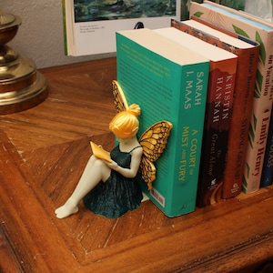 May include: A fairy ornament with golden wings and hair, reading a book, rests against a row of books. The books include titles such as "A Court of Mist and Fury" and "Throne of Glass". The scene is set on a wooden table.