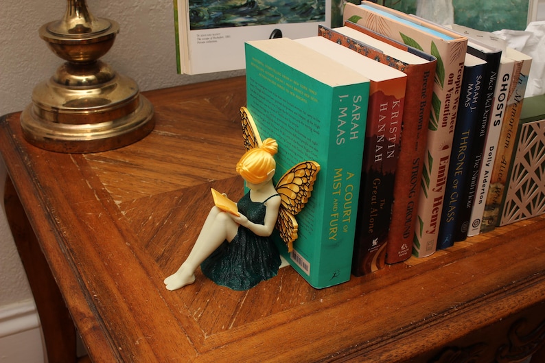 May include: A fairy ornament with golden wings and hair, reading a book, rests against a row of books. The books include titles such as "A Court of Mist and Fury" and "Throne of Glass". The scene is set on a wooden table.