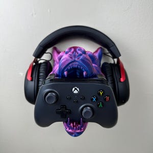 May include: A 3D-printed bulldog-shaped headphone stand in purple and blue, holding a black Xbox controller. Black and red headphones rest on the stand. The controller has colored buttons and a directional pad.