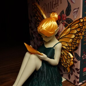May include: A fairy figurine with golden wings and hair, wearing a dark green dress, reading a book. The fairy is positioned next to a row of books, one of which has the title "Twilight". The fairy is holding an orange book.
