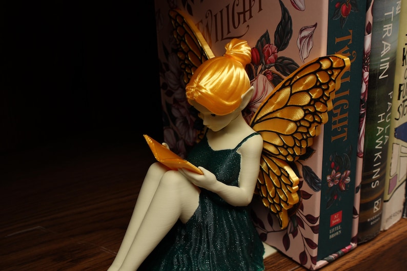 May include: A fairy figurine with golden wings and hair, wearing a dark green dress, reading a book. The fairy is positioned next to a row of books, one of which has the title "Twilight". The fairy is holding an orange book.