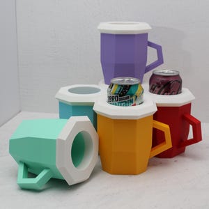 May include: A collection of five geometric, faceted beverage can holders with handles, featuring a white rim and base. The holders are colored in purple, light blue, mint green, yellow, and red, with two aluminum soda cans inserted inside.