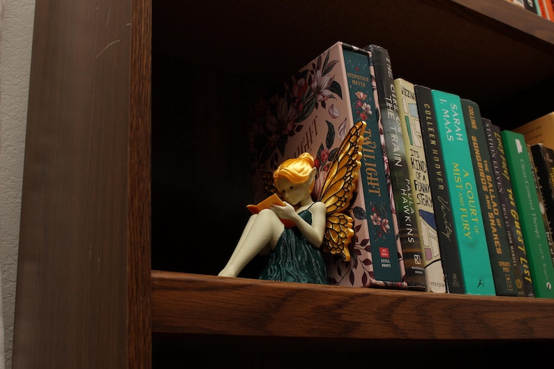 May include: A fairy figurine with golden wings and yellow hair is reading a book on a wooden shelf, in front of a row of books. The books include titles such as "Twilight" and "A Court of Mist and Fury".