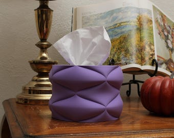 May include: A lavender tissue box with a geometric design, filled with white tissues. The box is on a wooden surface, next to a brass lamp and a decorative pumpkin. A book with landscape art is in the background.