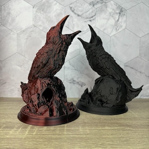 May include: Two 3D printed crow figurines with open beaks, one is black and the other is a dark red color. Both are perched on a skull-shaped base.