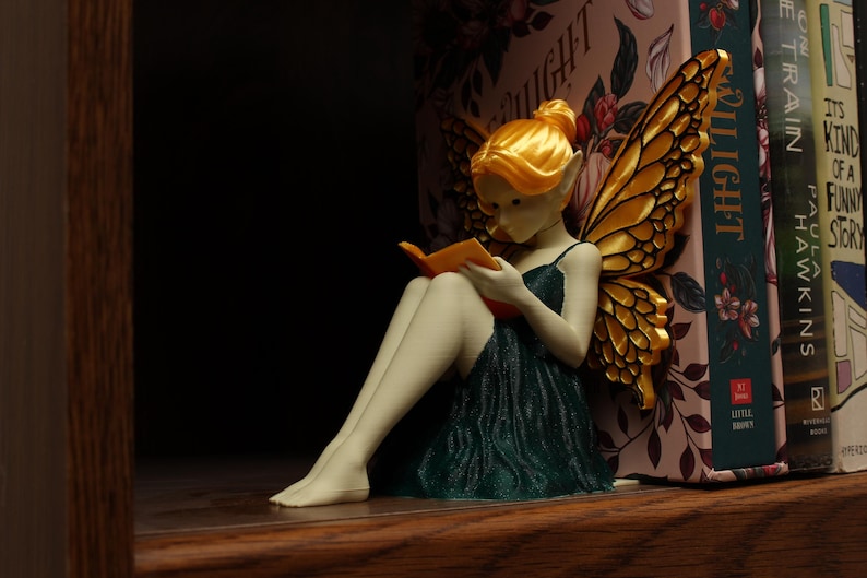 May include: A fairy figurine with golden wings and hair, reading a book, dressed in a dark green gown. The figurine is positioned on a wooden shelf beside a collection of books. The wings feature an intricate, textured pattern.