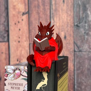 May include: A red dragon figurine reading a book, perched on top of a stack of books. The dragon has wings and is wearing a red cloak. The books have floral and dragon-themed covers.