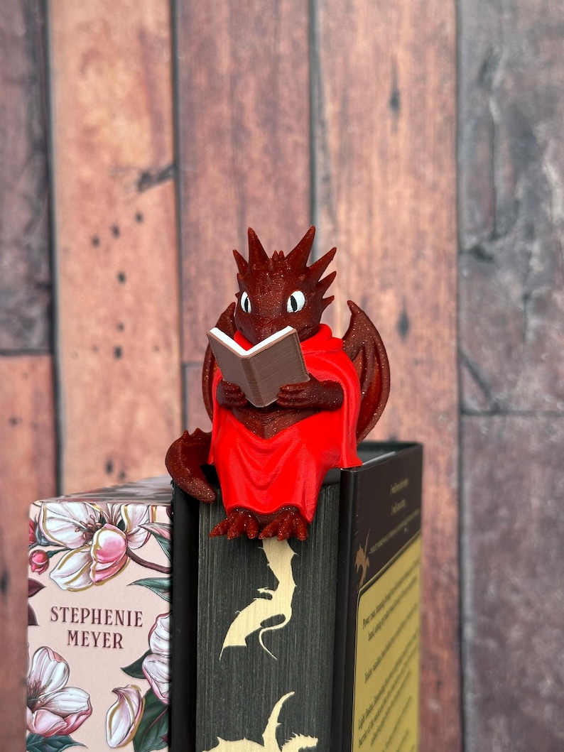 May include: A red dragon figurine reading a book, perched on top of a stack of books. The dragon has wings and is wearing a red cloak. The books have floral and dragon-themed covers.