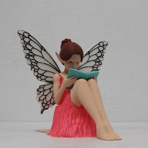 May include: A fairy figurine with large, detailed wings, wearing a pink dress, and reading a teal book. The fairy has brown hair and pointed ears. The wings are white with black detailing. The fairy is sitting down.