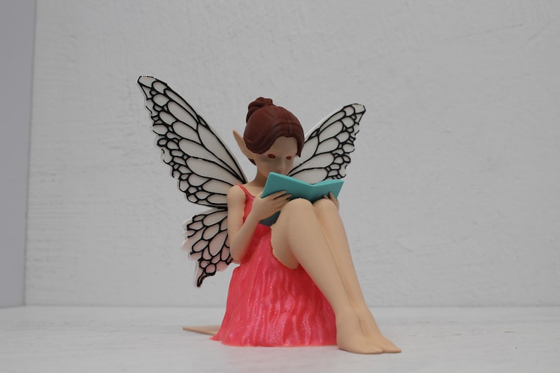 May include: A fairy figurine with large, detailed wings, wearing a pink dress, and reading a teal book. The fairy has brown hair and pointed ears. The wings are white with black detailing. The fairy is sitting down.