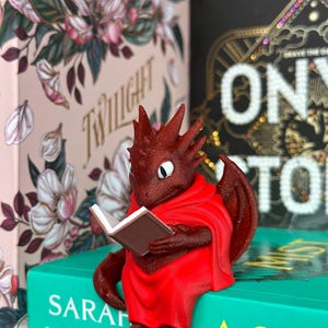 May include: A red dragon figurine wearing a red cloak, reading a small book. The dragon is perched on a teal book with white text. Other books with floral designs and gold lettering are in the background.