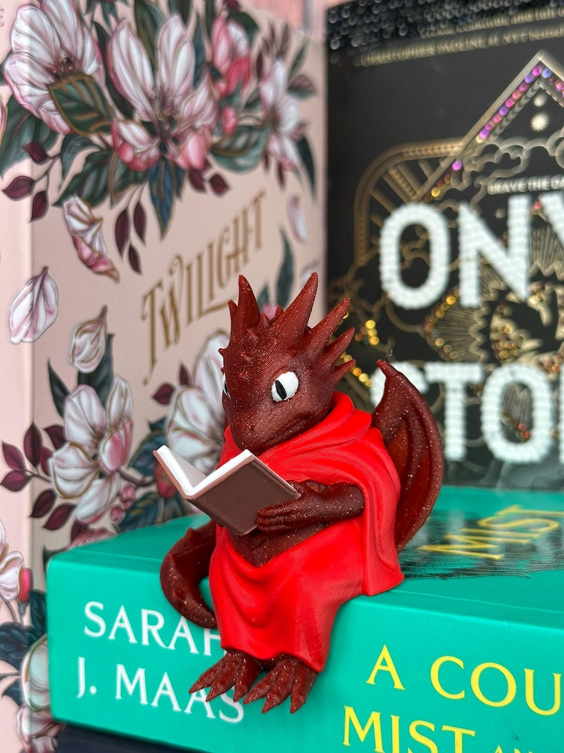 May include: A red dragon figurine wearing a red cloak, reading a small book. The dragon is perched on a teal book with white text. Other books with floral designs and gold lettering are in the background.