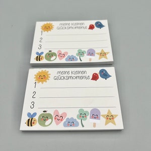 Könnte beinhalten: Zwei weiße Notizblock-Sets mit einem farbenfrohen Design. Jedes Set hat eine Sonne, einen Vogel, eine Biene, einen Apfel, ein Herz, ein Kleeblatt, ein Eis am Stiel und einen Stern. Der Text auf dem Notizblock lautet "meine kleinen Glücksmomente", was übersetzt "meine kleinen Glücksmomente" bedeutet.