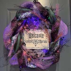 May include: A Halloween wreath with purple, black, and green mesh ribbon, adorned with small lights and sprigs. A wooden sign in the center reads "Welcome Foolish Mortals" in a gothic font, creating a spooky, festive decoration.