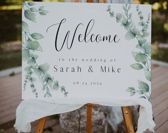 May include: A rectangular white wedding welcome sign on a wooden easel, featuring green watercolor eucalyptus leaf illustrations on the sides and black text reading Welcome to the wedding of Sarah & Mike 08.24.2024.