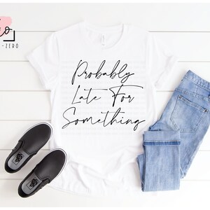May include: White t-shirt with black text that reads "Probably Late For Something" in a cursive font. The shirt is displayed on a white wooden surface with a pair of black slip-on shoes and blue jeans.