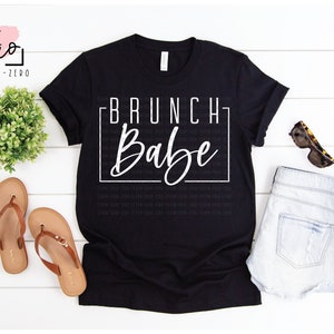 May include: Black t-shirt with white text that reads "BRUNCH Babe" in a square frame. The shirt is on a white wooden surface with a pair of brown sandals, a pair of denim shorts, and sunglasses.