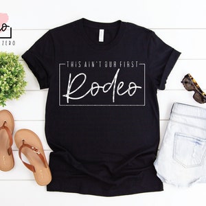 Puede incluir: Camiseta negra con texto blanco que dice "This ain't our first Rodeo".