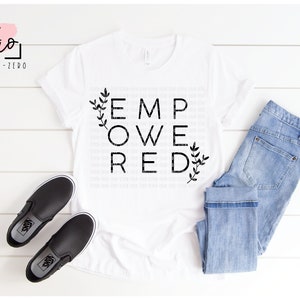 Può includere: Una maglietta bianca con la parola "EMPOWERED" stampata in lettere nere con piccoli disegni floreali su entrambi i lati. La maglietta è stesa su una superficie di legno bianco con un paio di scarpe nere da barca e un paio di jeans blu.