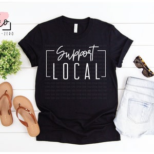 May include: Black t-shirt with white text that reads "Support Local" in a square frame. The shirt is laying flat on a white wooden surface with a pair of brown sandals, a pair of denim shorts, and sunglasses.