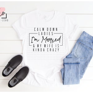 May include: White t-shirt with black text that reads "Calm down ladies I'm married & my wife is kinda crazy". The shirt is displayed on a white wooden surface with a pair of black slip-on shoes and blue jeans.