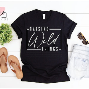 May include: Black t-shirt with white text that reads "Raising Wild Things" in a box. The shirt is on a white wood background with a pair of brown sandals, a pair of denim shorts, and sunglasses.