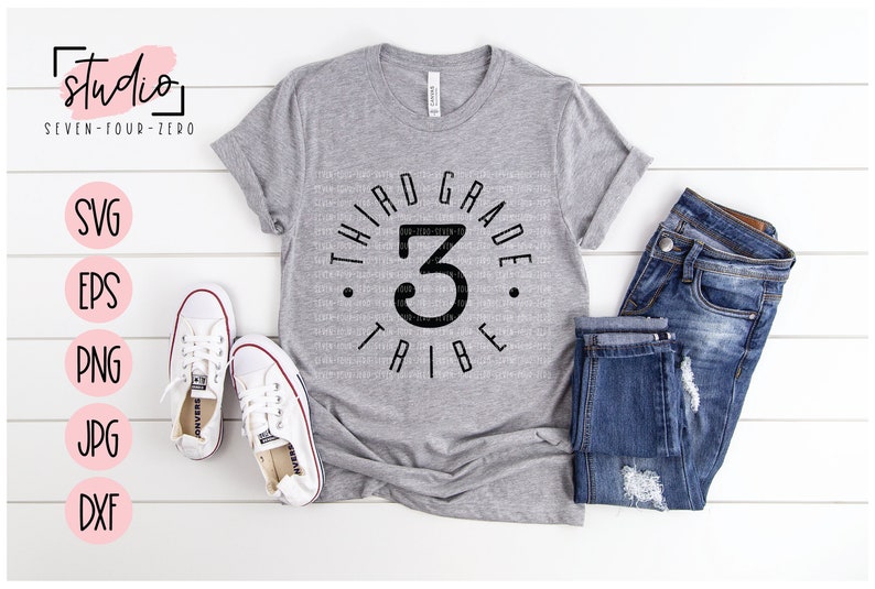 May include: A gray t-shirt with a black circle design that says "Third Grade Tribe" with the number 3 in the center. The shirt is laid out on a white wooden surface with a pair of white sneakers and blue jeans.
