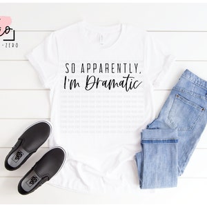 Könnte beinhalten: Weißes T-Shirt mit dem Text "SO APPARENTLY, I'm Dramatic" in schwarzer Schrift. Das Shirt wird auf einer weißen Holzoberfläche mit einem Paar schwarzer Slipper und einer blauen Jeans gezeigt.