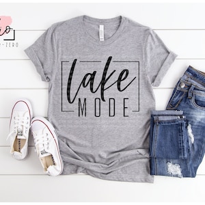 May include: A heather gray t-shirt with the words "Lake Mode" in a black, stylized font. The shirt is paired with blue jeans and white canvas sneakers. The image also includes design file format options.