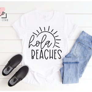 Op de afbeelding: Wit T-shirt met zwarte tekst die "hola beaches" zegt en een zon-graphic. Het shirt ligt op een wit houten oppervlak met een paar zwarte slip-on schoenen en een blauwe jeans.