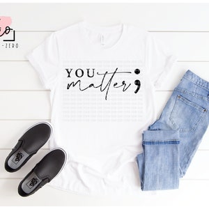 May include: White t-shirt with the text "YOU matter" in black script with a semicolon at the end. The shirt is laying flat on a white wooden surface with a pair of black slip-on shoes and blue jeans.