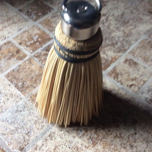 Puede incluir: Un pequeño cepillo de barrer de mano con una parte superior plateada y un mango. La escoba está hecha de fibras naturales y tiene una base de tejido marrón.