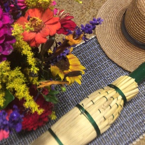 Puede incluir: Un primer plano de una escoba hecha a mano con un mango verde y cerdas de paja natural. La escoba está sobre una alfombra tejida azul y blanca con un ramo de flores de colores y un sombrero de paja en el fondo.