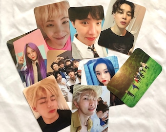 May include: A collection of photo cards featuring various K-pop idols. The cards are printed on white cardstock and feature different images of the idols, including close-ups, group shots, and performance shots. The cards are all in portrait orientation and have a white border around the image.