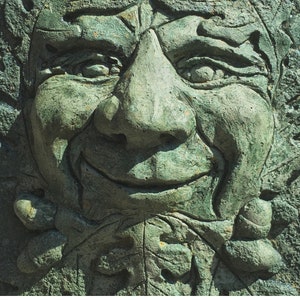 Op de afbeelding: Een close-up van een groene stenen sculptuur van een gezicht met een grote neus en een brede glimlach. De sculptuur is gedetailleerd met rimpels en lijnen rond de ogen en de mond.