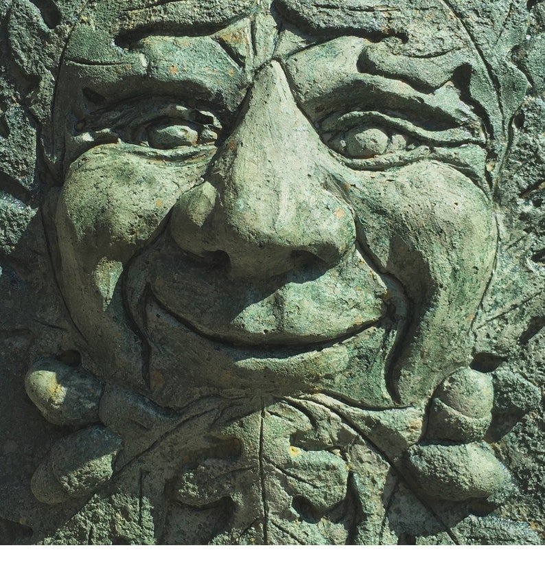 Op de afbeelding: Een close-up van een groene stenen sculptuur van een gezicht met een grote neus en een brede glimlach. De sculptuur is gedetailleerd met rimpels en lijnen rond de ogen en de mond.