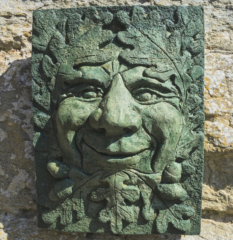 Op de afbeelding: Een groene stenen sculptuur van een gezicht met bladeren en eikels eromheen. Het gezicht glimlacht en heeft een grote neus.